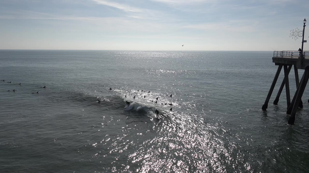 At Huntington Beach, whitewater starts to spill across a wave as multiple surfers sit or paddle nearby, framed by the dark pier supports on the right.