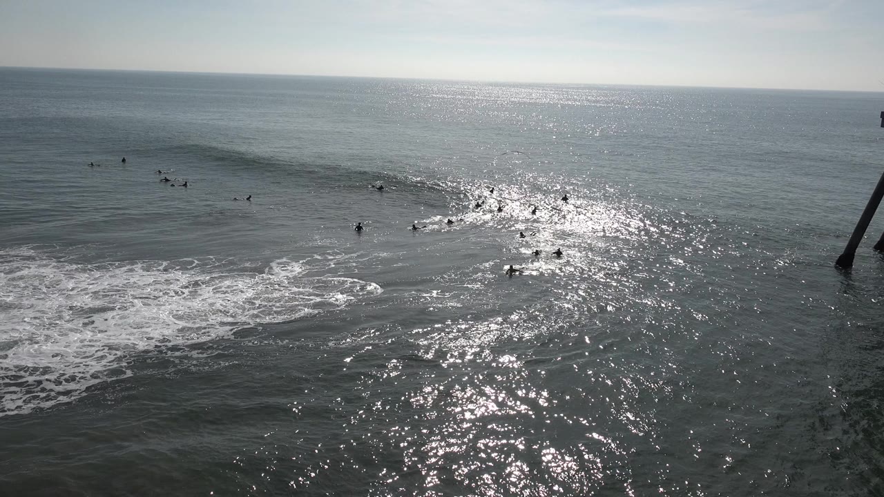 Distant surfers wait in the water at Huntington Beach, California as a low wave rises beside white foam and silver glare spreads across the ocean.