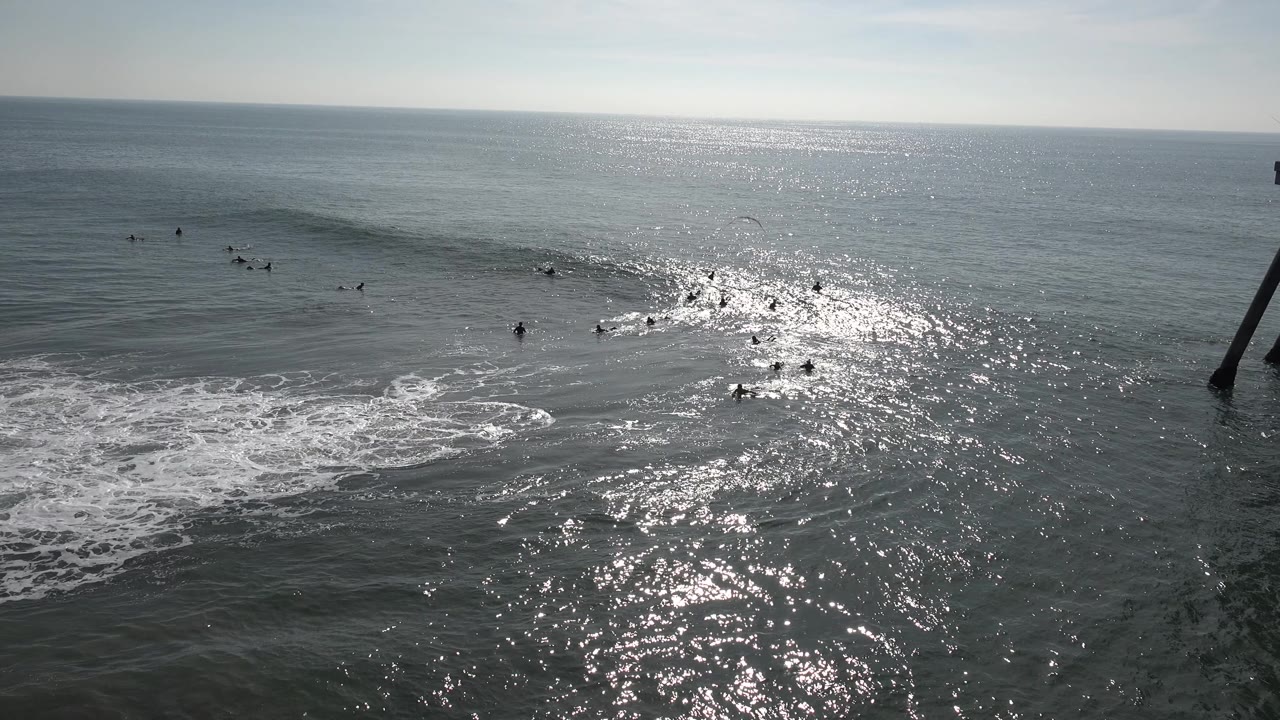 Surfers remain scattered beyond the foam at Huntington Beach, California while the wave line softens and sunlight reflects in a wide patch across the water.