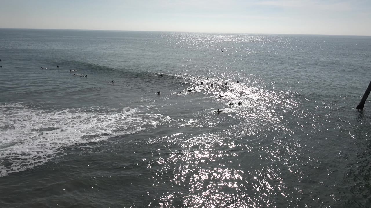 At Huntington Beach, California, distant surfers cluster around a developing wave while foam spreads across the lower frame and a dark post-like structure sits near the horizon line.