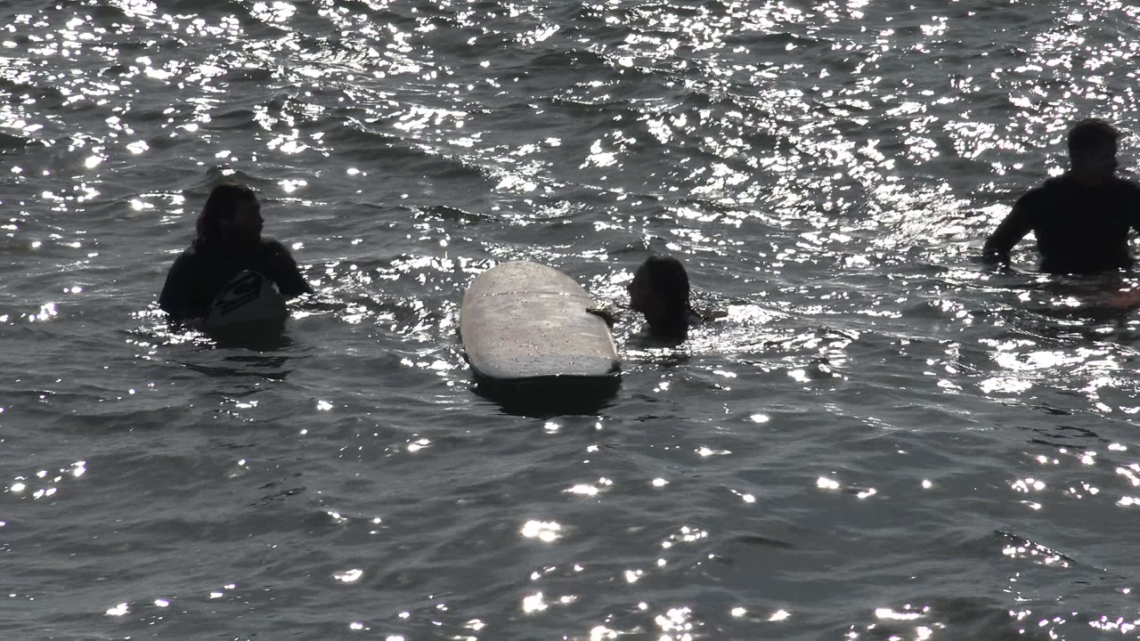 At Huntington Beach, several people sit or float in the ocean near a pale surfboard as bright sunlight reflects across the water.