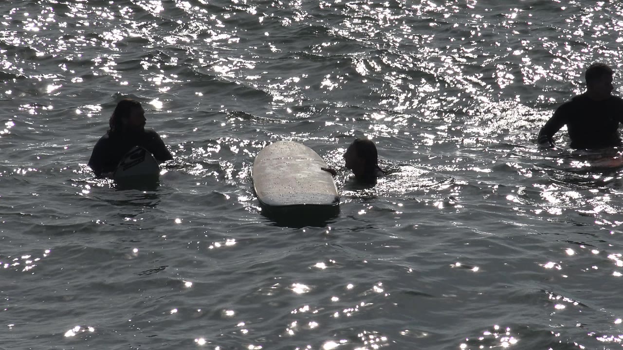 At Huntington Beach, the surfboard floats between two people while other swimmers appear farther back in heavily backlit ocean water.