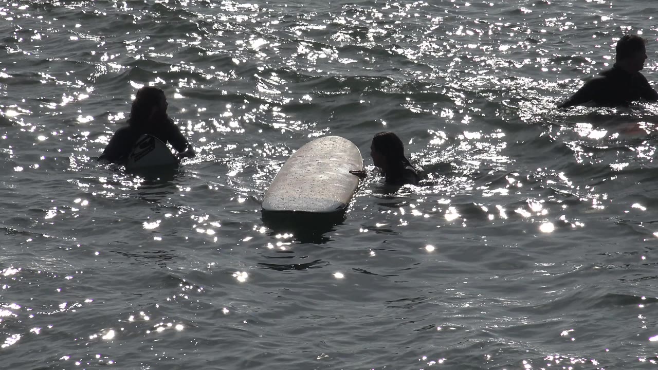 At Huntington Beach, silhouetted people float around a pale surfboard as the wave texture shifts and sun flecks fill the background.