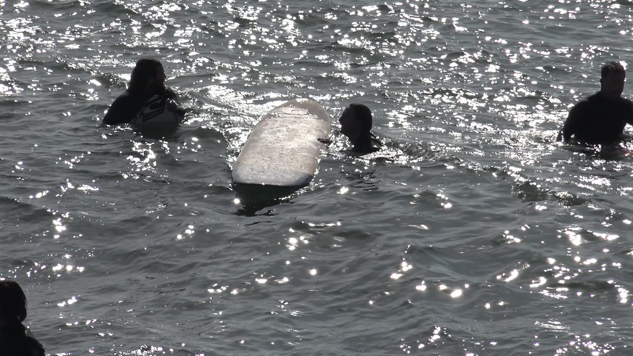 A surfboard floats beside people in choppy sunlit water.