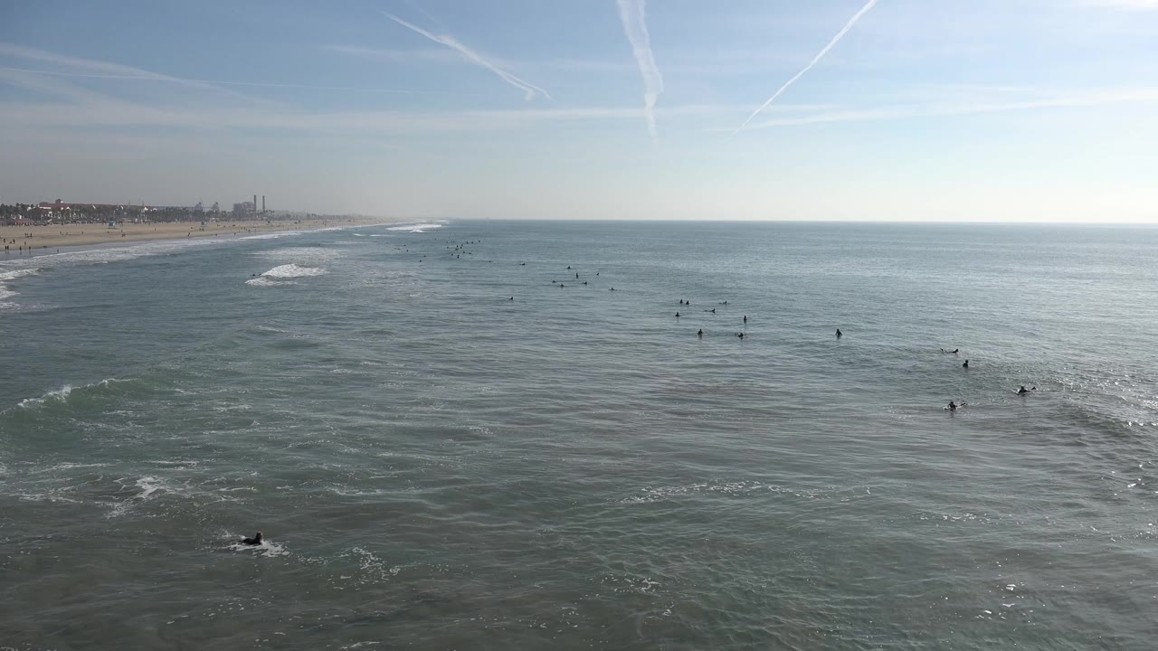 Wide view of Huntington Beach ocean with small surfers spread across the water, low waves forming, sandy beach at left, and bright sky above.