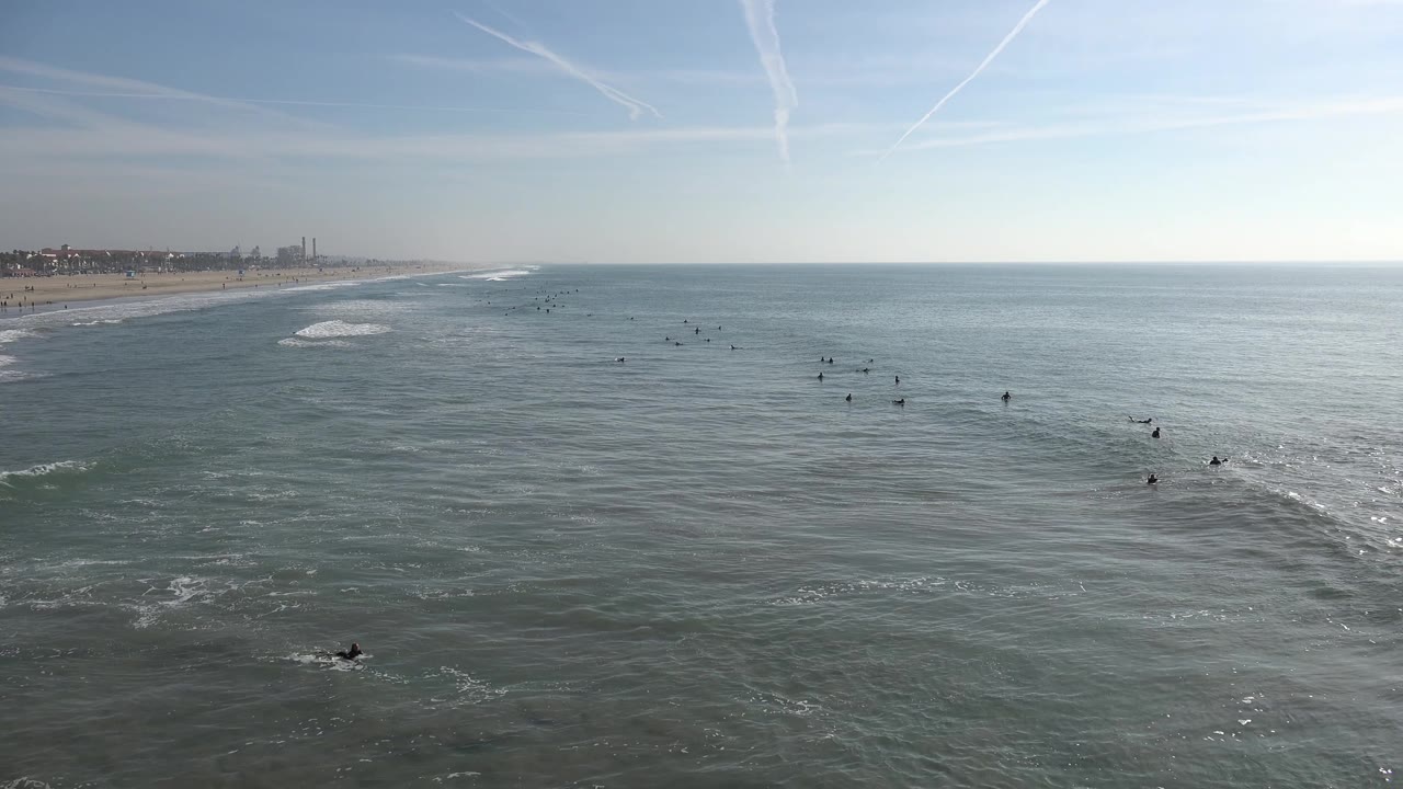 A wide Huntington Beach frame shows blue-gray ocean, distant surfers clustered near the center and right, a thin surf line near shore, and a clear horizon.