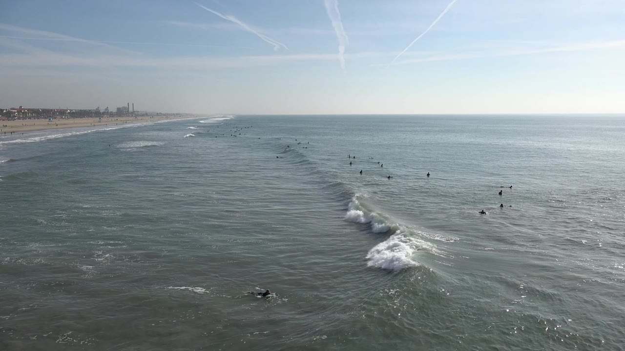 Wide view of Huntington Beach ocean with small surfers and swimmers spaced across the water, a sandy coastline at left, and thin white waves under a pale blue sky.