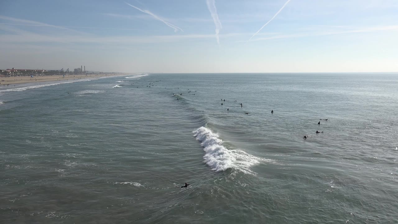 A white wave breaks in the midground off Huntington Beach while small dark figures wait farther out and the shoreline remains visible along the left edge.