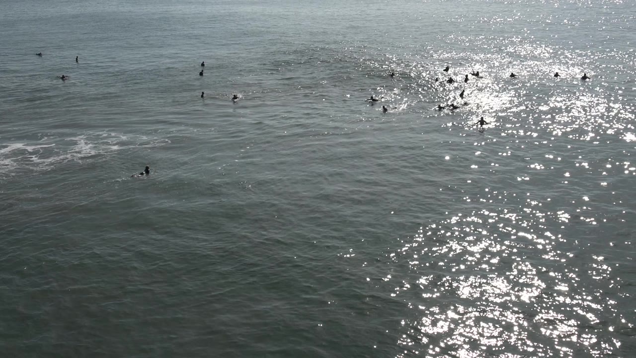 Distant surfers and swimmers are spread across gray green ocean water in Huntington Beach, with a strong path of sun glare near the center.