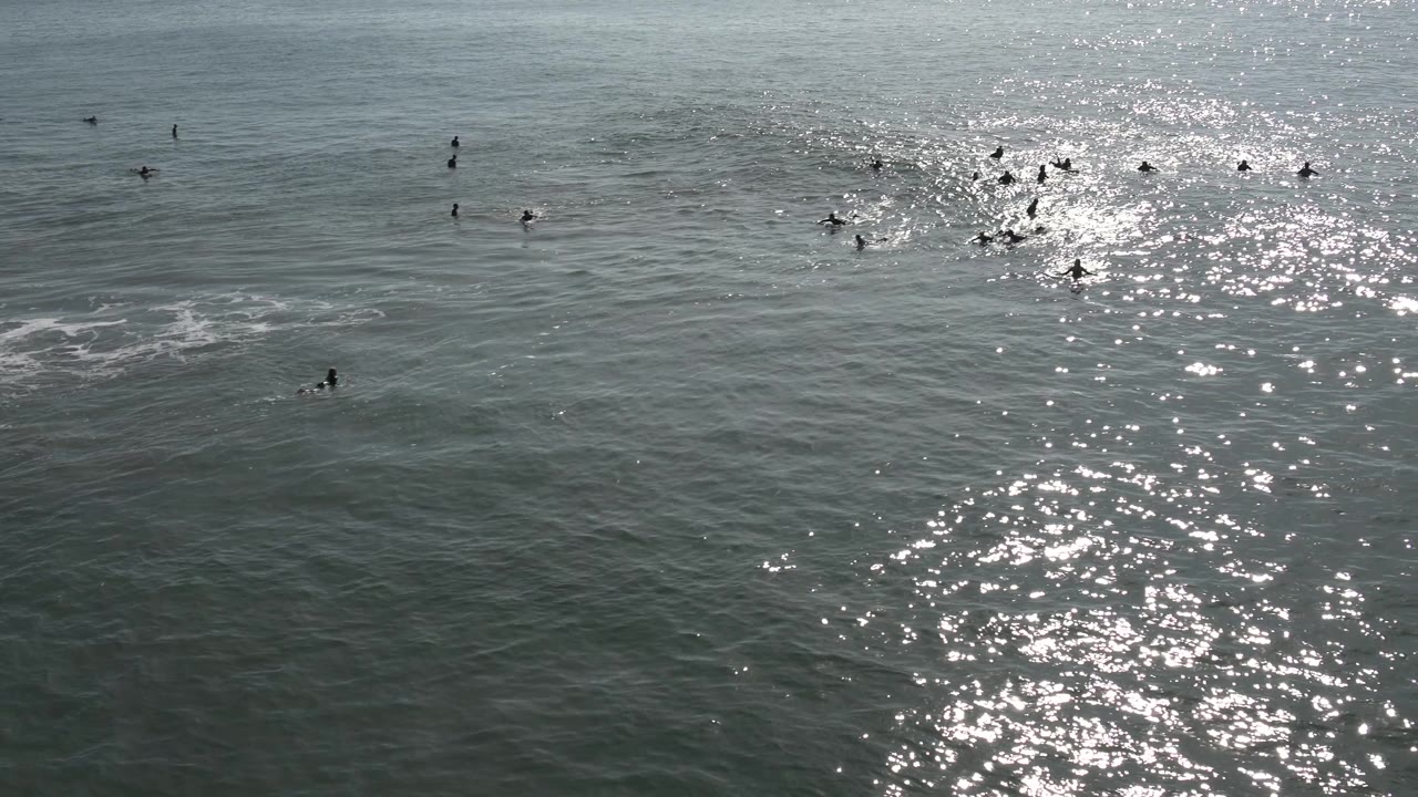 A bright vertical reflection crosses the Huntington Beach water while small dark surfer silhouettes wait beyond a low wave.