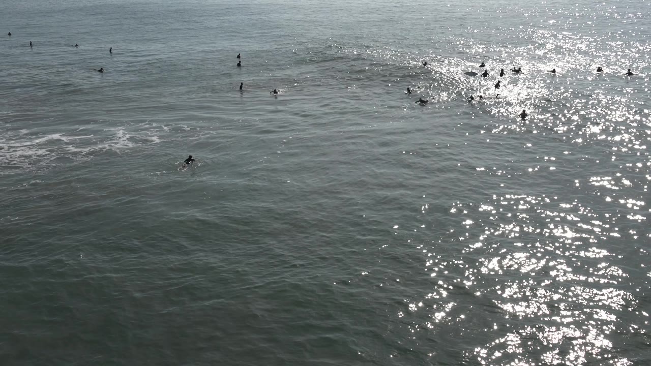 A small wave lifts across the right side of the Huntington Beach ocean scene as distant surfers cluster near the glittering sunlight.