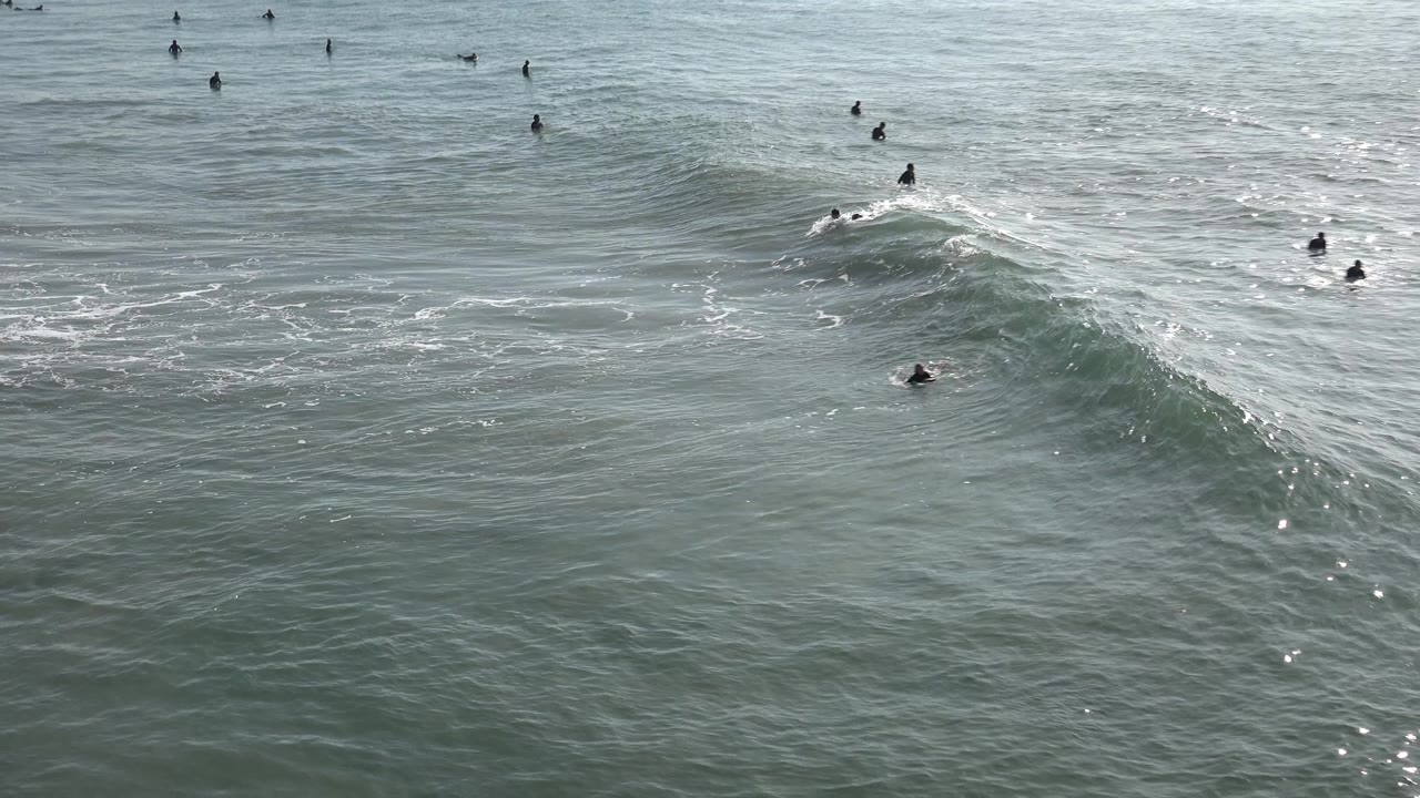Wide Huntington Beach ocean view with a large gray-green swell rising across the frame and small swimmers or surfers scattered in the water.
