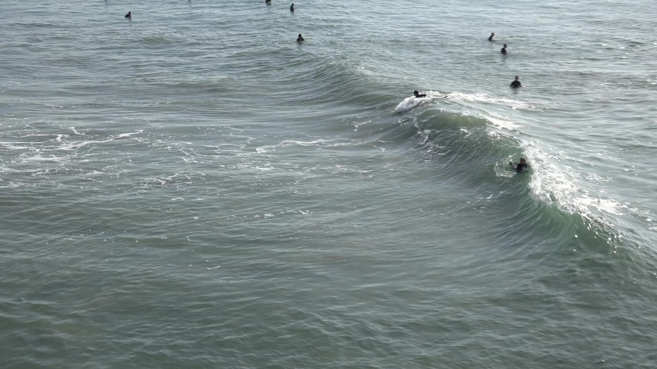 Wide Huntington Beach water view with a swell breaking at right and multiple small figures visible beyond and beside the wave.