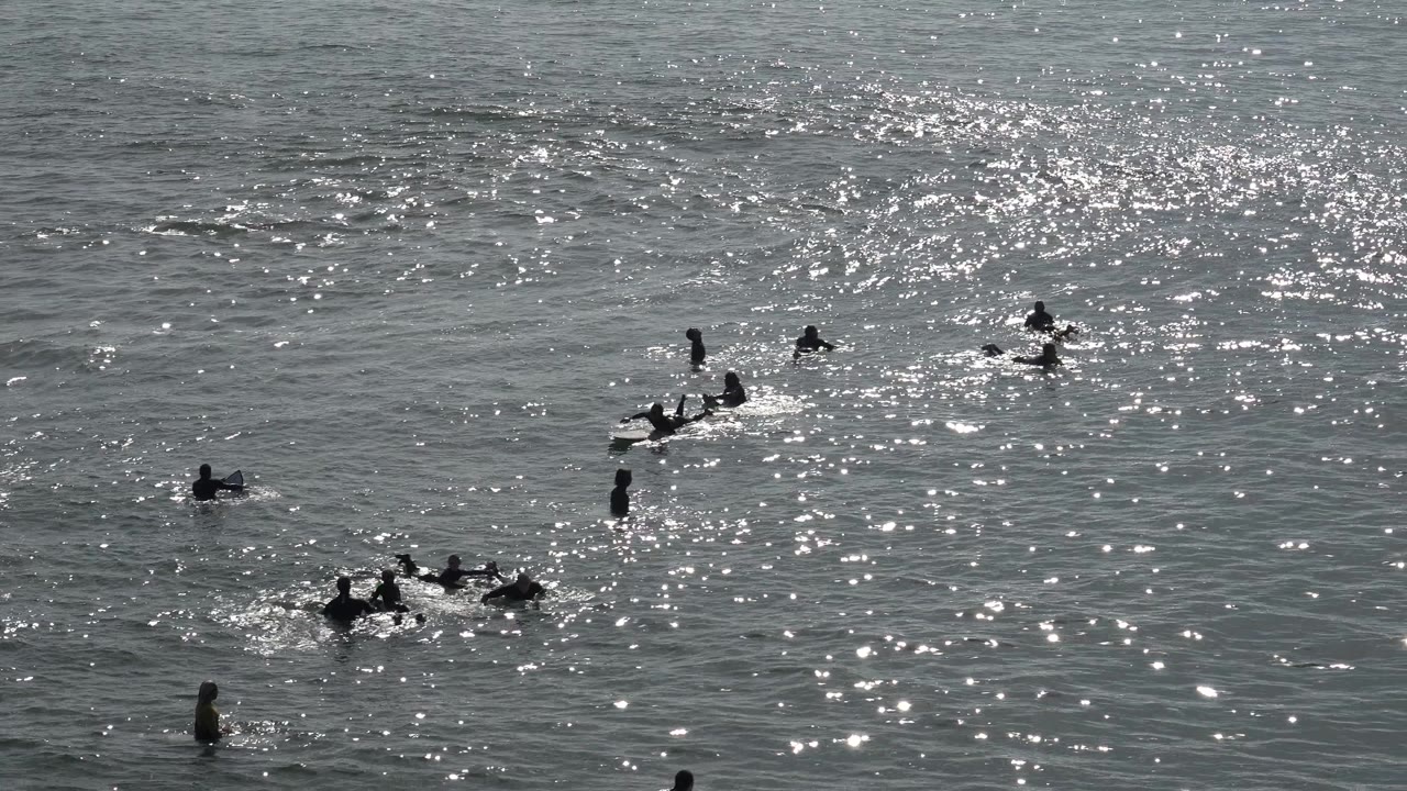 At Huntington Beach, swimmers and board riders are spread across gray-green water with bright sun glare across the surface near the start of the clip.