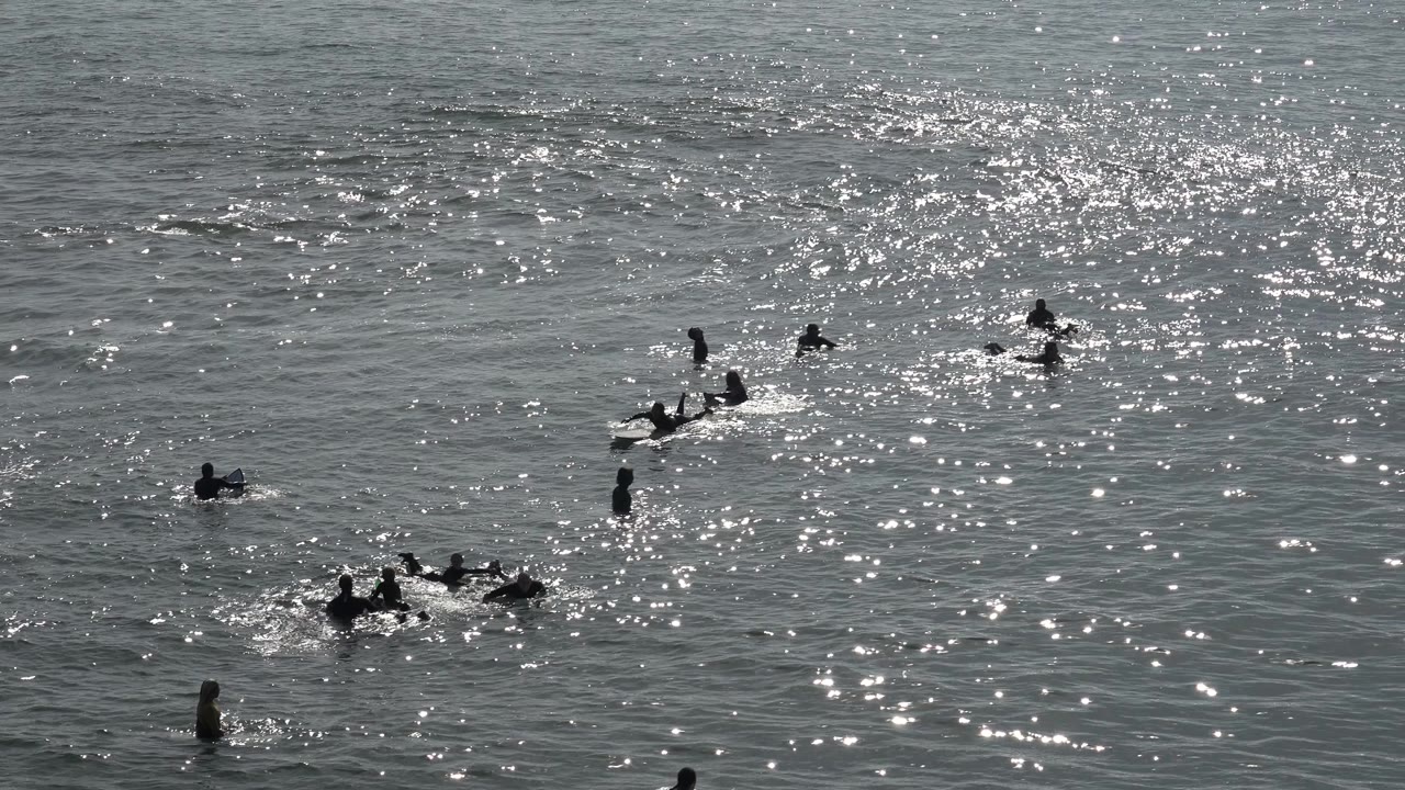 A surfer paddles among swimmers in glittering water.