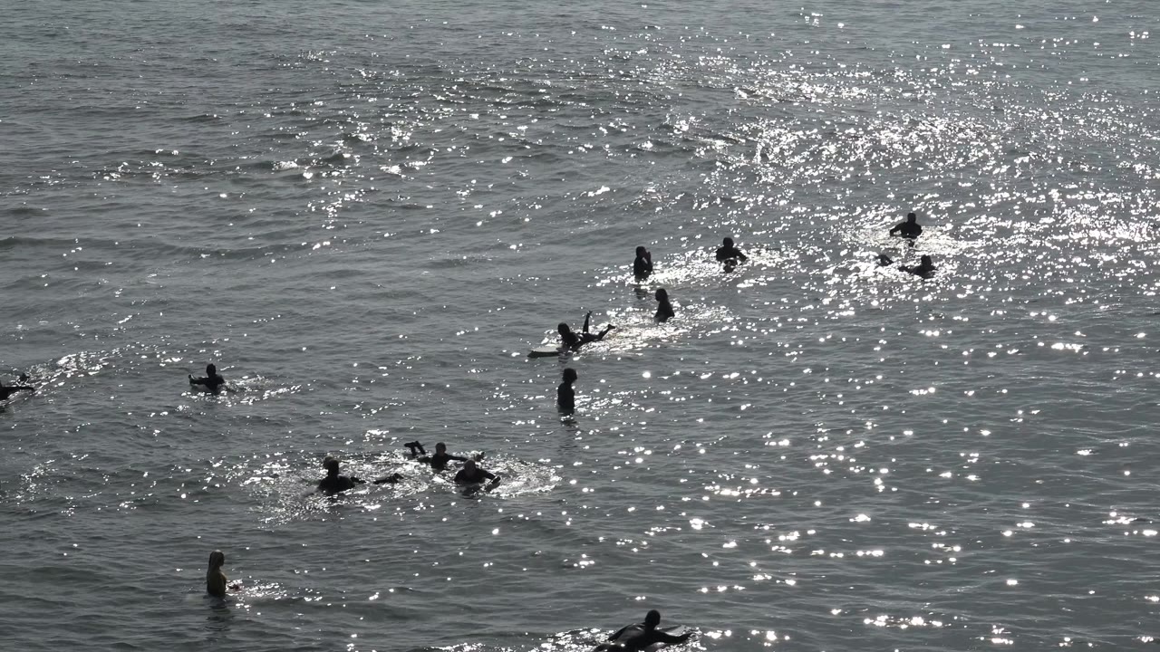 At Huntington Beach, several people cluster around a small wave while other swimmers and board riders remain spread farther back in the water.