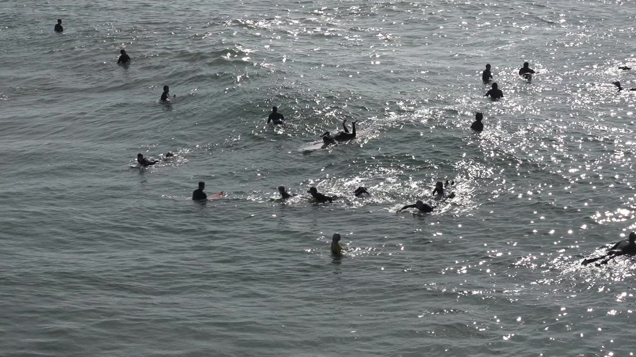 Surfers and swimmers are spread across reflective ocean water at Huntington Beach, California, with small waves forming around them at the start of the clip.