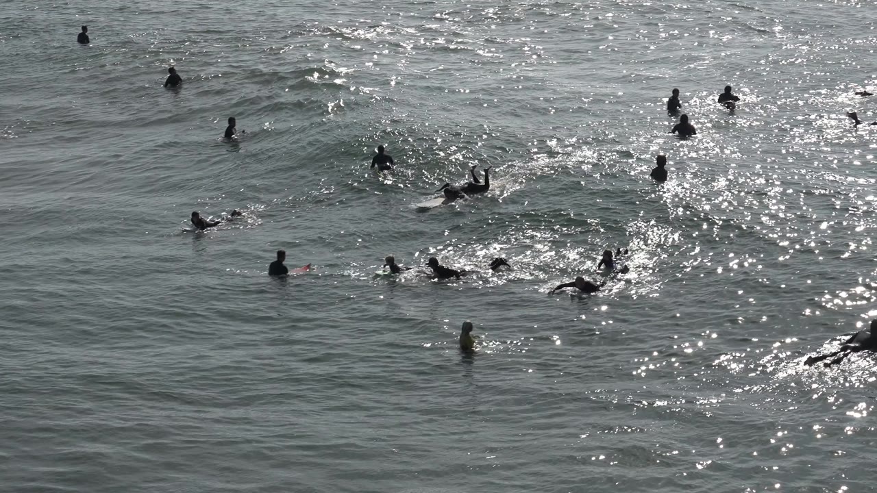 People in dark swimwear and wetsuits float and paddle through gray-green water at Huntington Beach, California, as sunlight reflects across the midground.