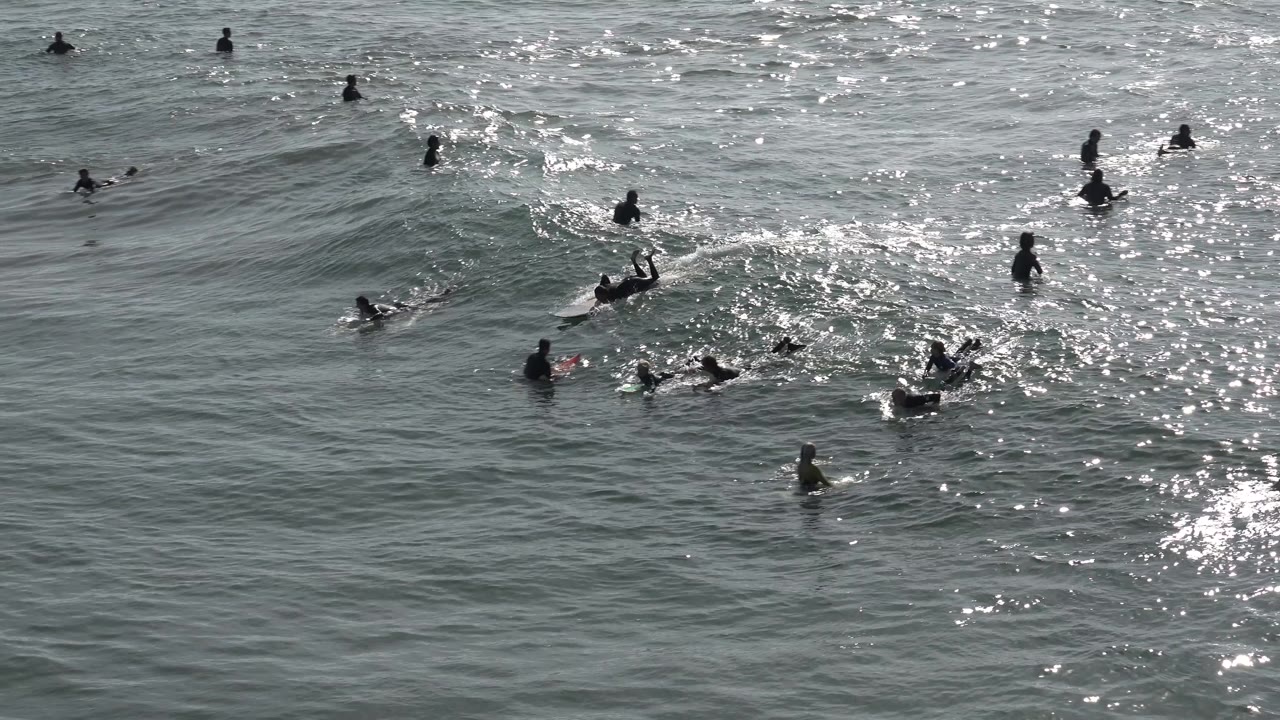 A cluster of surfers and swimmers gathers near a small breaking wave at Huntington Beach, California, with bright water glare behind them.