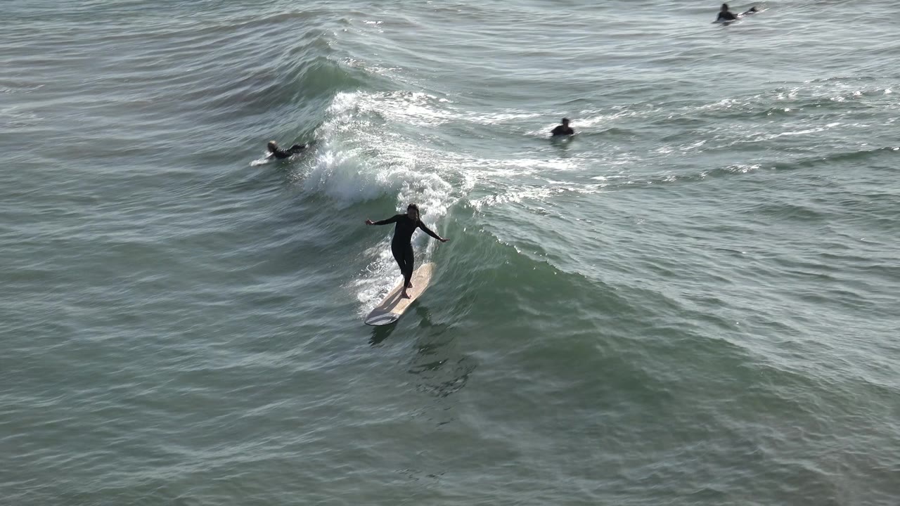 A surfer in a black wetsuit rides a longboard across green-gray water at Huntington Beach, California, with whitewater forming behind and other surfers farther out.