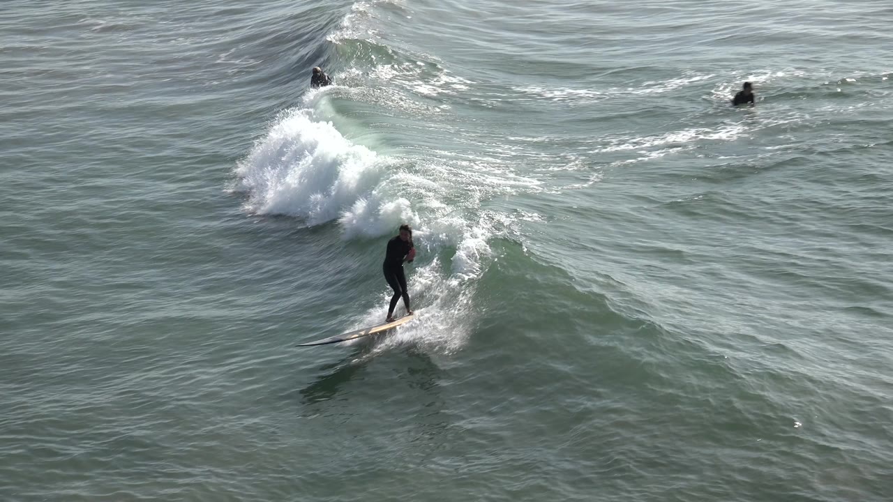 A surfer in a black wetsuit rides ahead of a stronger section of whitewater at Huntington Beach, California, with textured ocean surface filling the frame.