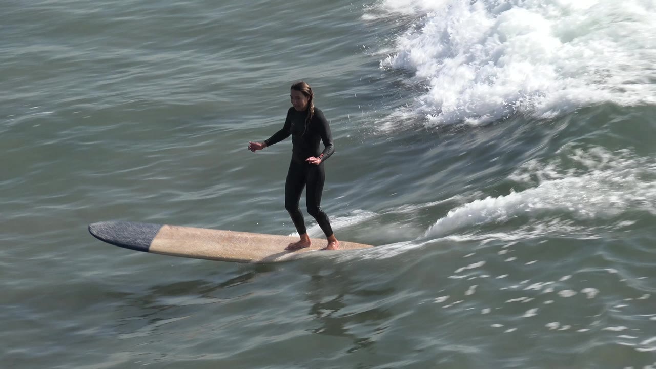 A young surfer in a black wetsuit rides a longboard across green-gray water at Huntington Beach, California, with white foam rising behind the board.