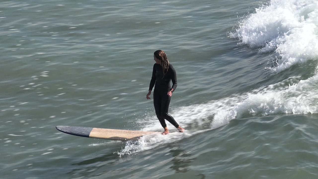 A wetsuited surfer rides ahead of whitewater at Huntington Beach, California, with both feet visible on the longboard and spray touching the rear of the board.