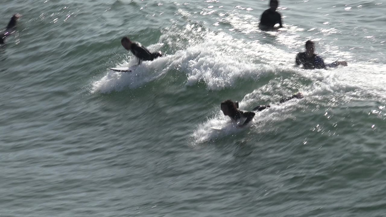 Surfer begins riding a foamy wave while others paddle nearby.