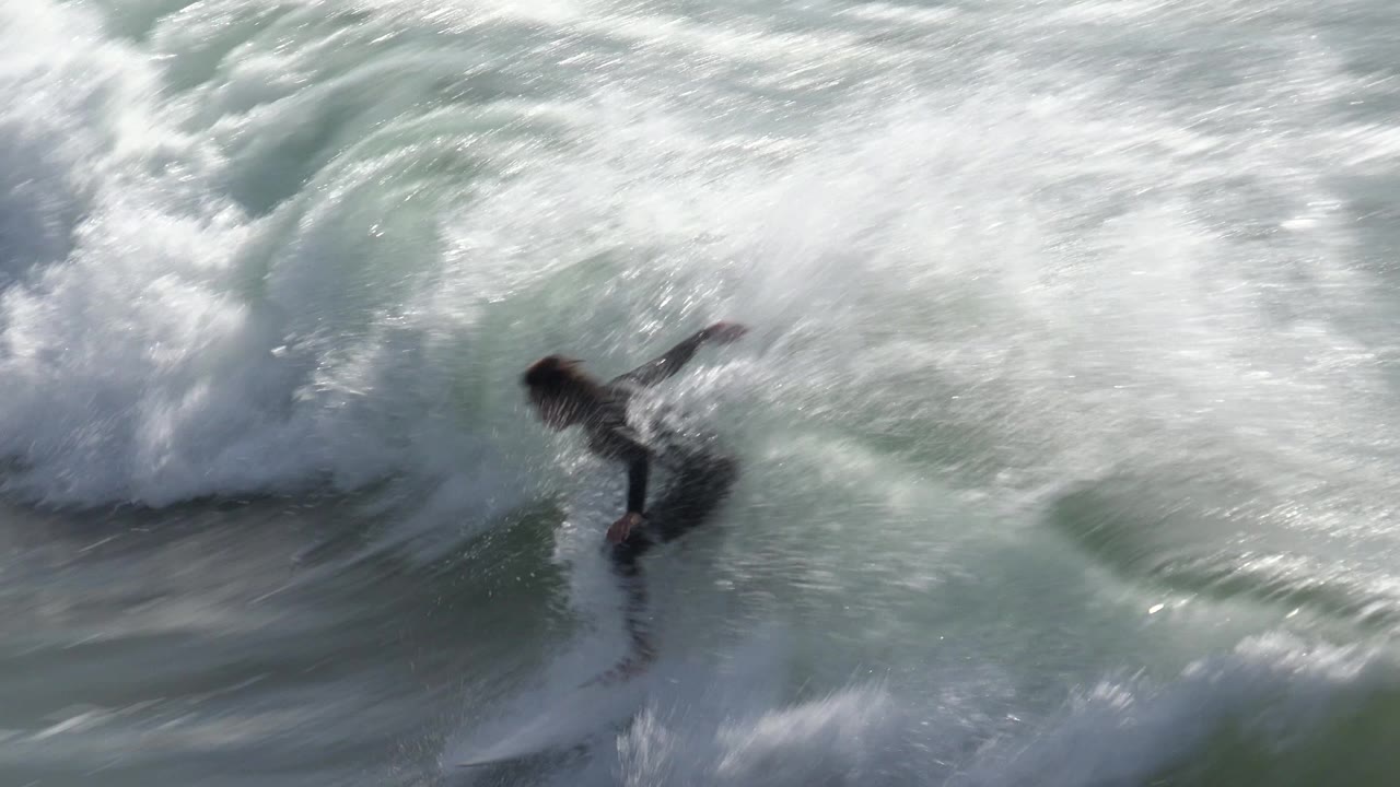 A surfer in a black wetsuit crouches low on a white board beneath breaking whitewater at Huntington Beach, California.