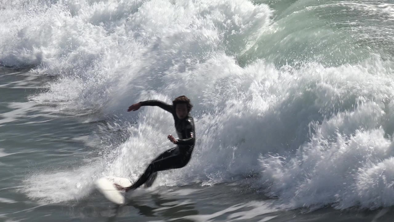A surfer stands on a white board with one arm extended as bright white surf breaks close behind at Huntington Beach, California.
