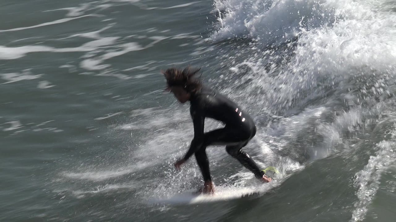 A woman in a black wetsuit rides a white surfboard on a breaking wave at Huntington Beach, California, with white spray rising behind her.