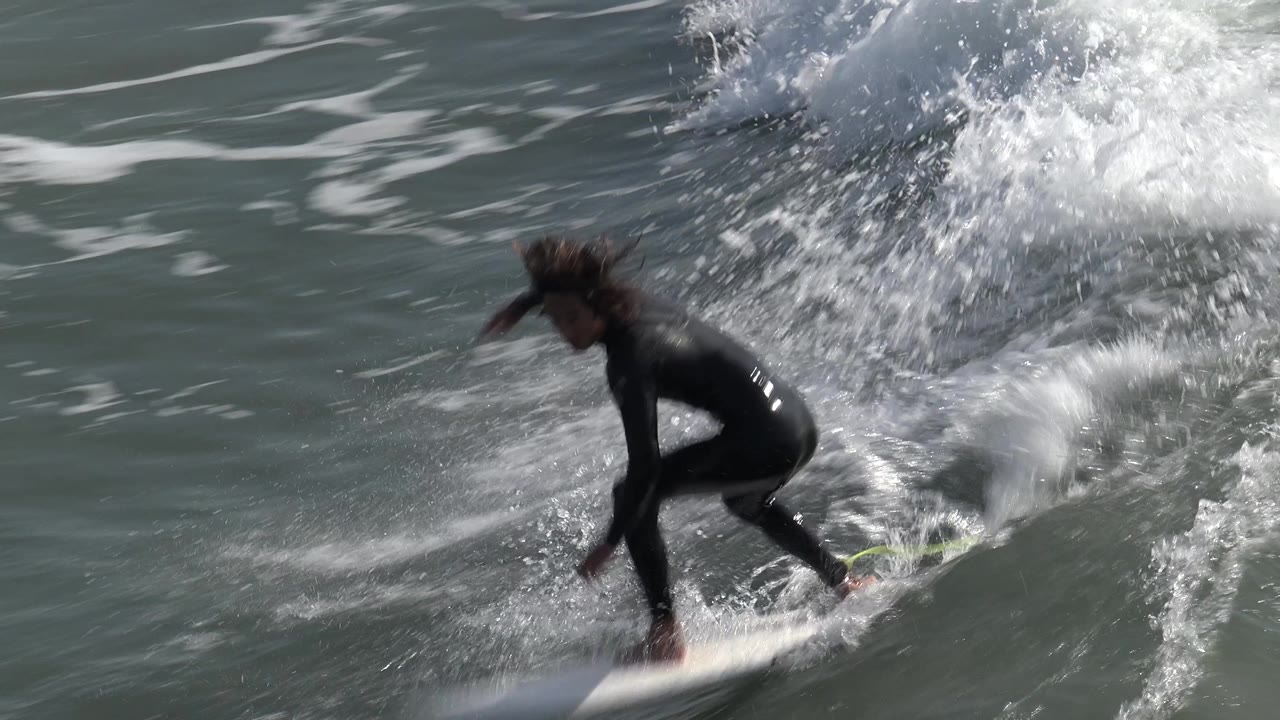 The surfer stays crouched on the white board at Huntington Beach, California, while the wave steepens and a fan of spray trails behind her.