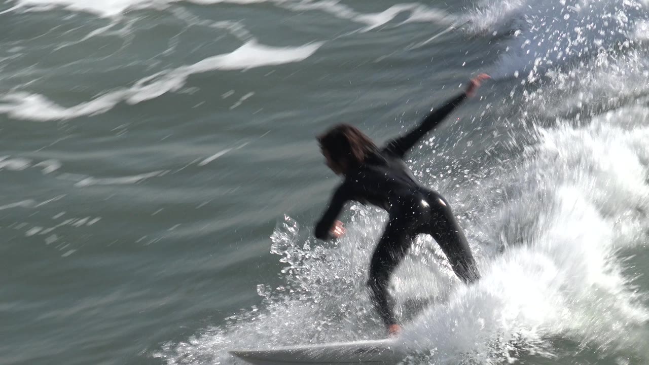 A woman in a black wetsuit leans over the surfboard at Huntington Beach, California, as whitewater breaks around the board and obscures part of the wave.
