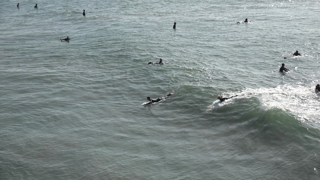 A surfer in a dark wetsuit rides along the right side of a breaking Huntington Beach wave, with several other surfers farther out across the green-gray water.