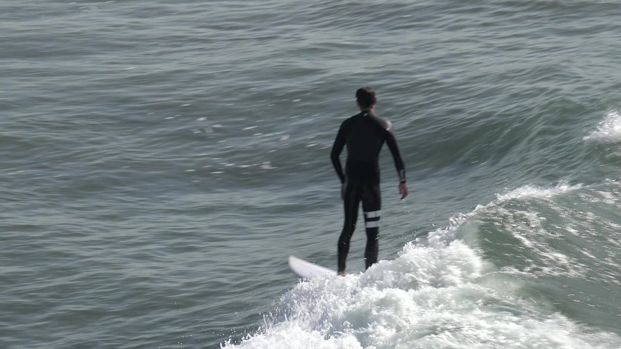 A surfer in a black wetsuit stands on a white surfboard over whitewater at Huntington Beach, California, with gray-green waves filling the frame.