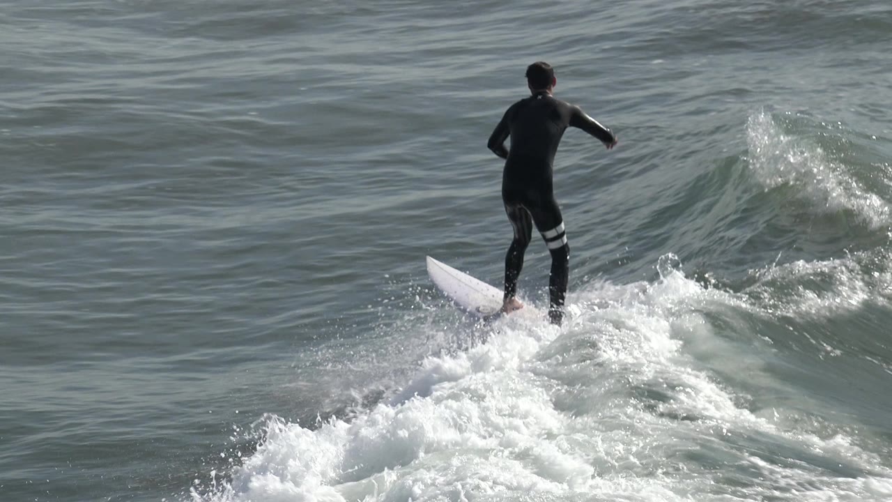 A wetsuited surfer rides over churning whitewater at Huntington Beach, California, with the white surfboard angled across the foamy wave.