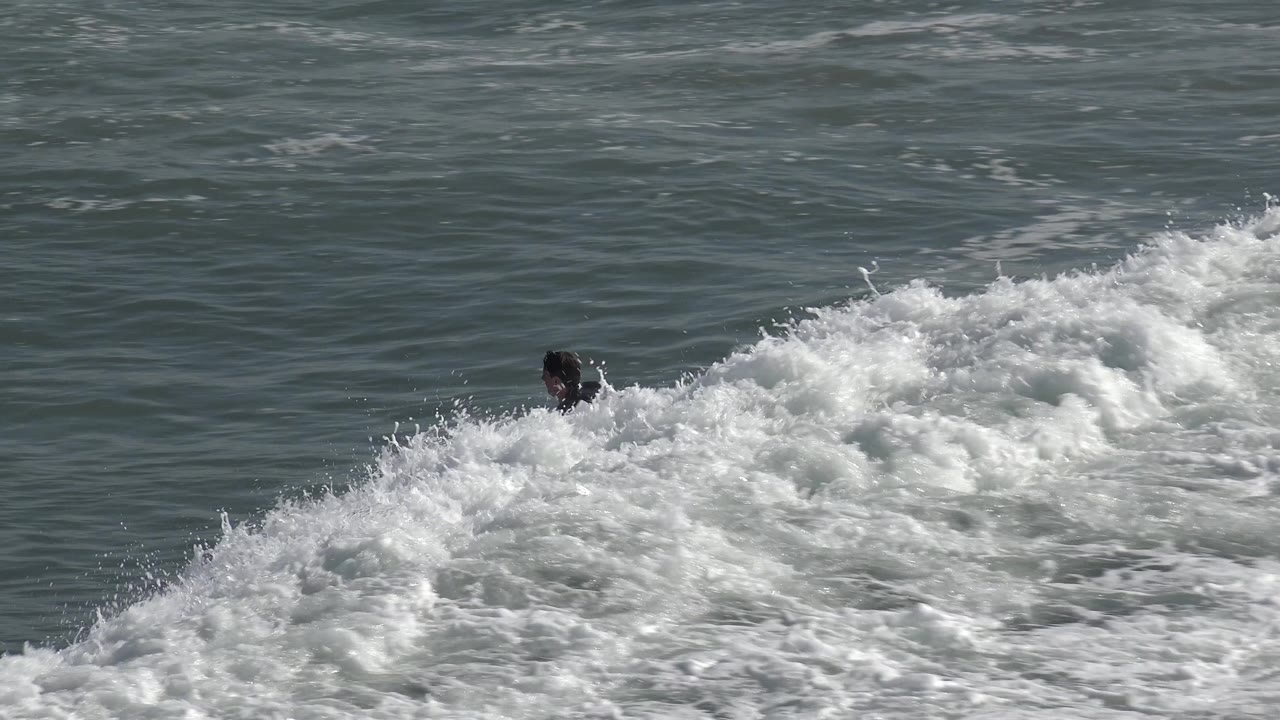 At Huntington Beach, California, a surfer in a dark wetsuit rides near the right edge of a broad whitewater section, with textured ocean behind him.