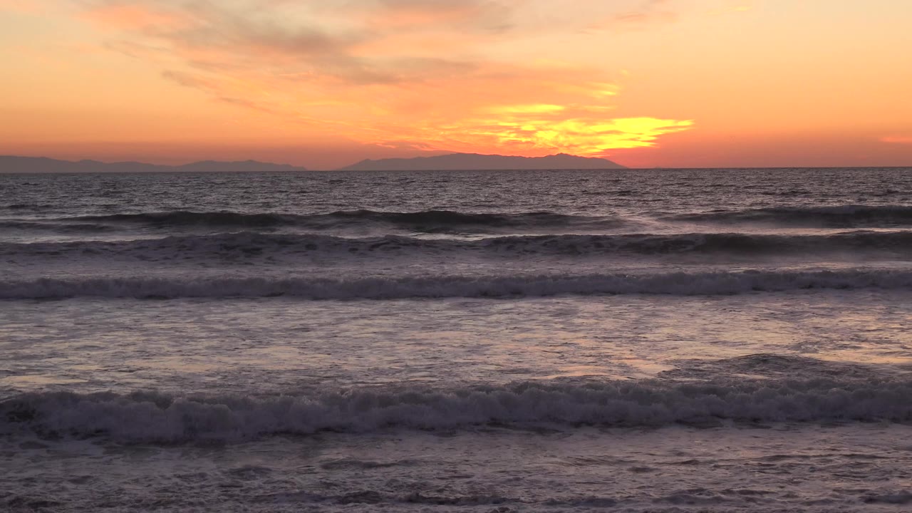 Huntington Beach ocean waves roll toward the camera beneath an orange sunset sky, with a dark horizon silhouette and no visible people.