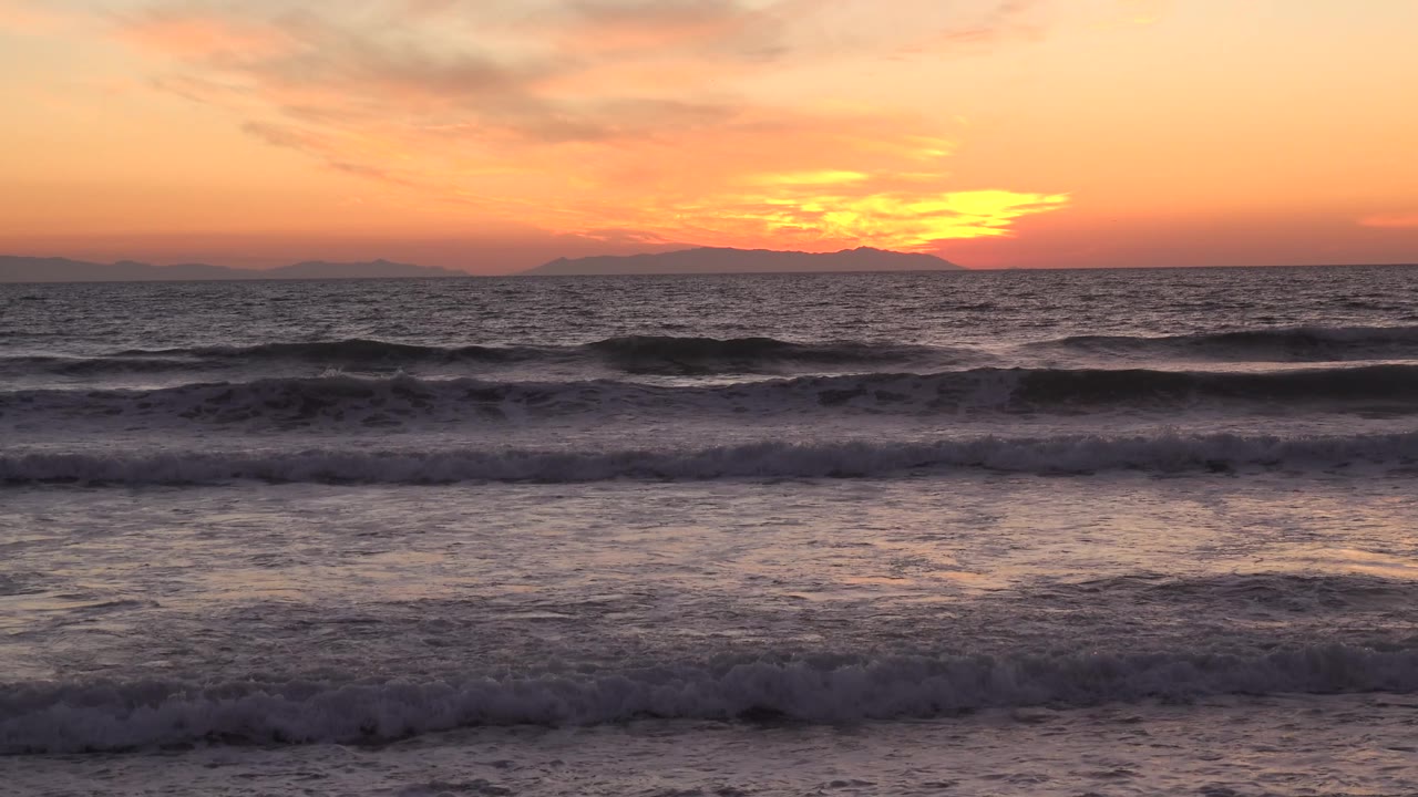 Huntington Beach waves break across the midground while the sunset glow sits behind a low dark landform, with no visible people.