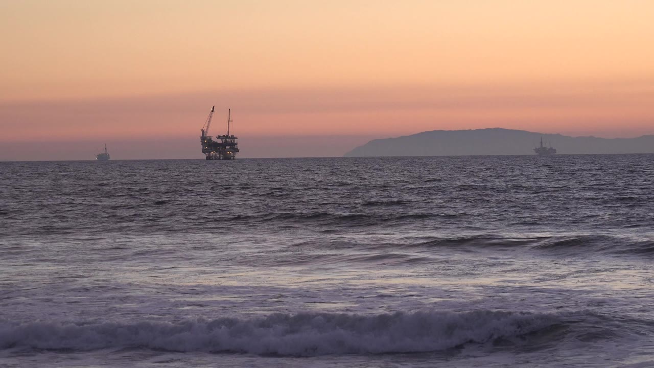 Opening frame of Huntington Beach twilight surf with low foreground waves, distant offshore structures, hazy hills, and no visible people.