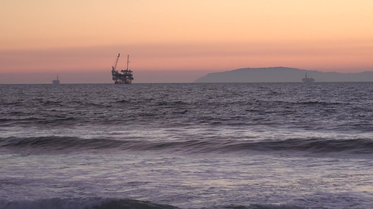 At Huntington Beach, offshore boats sit on the horizon while rough surf rolls through the foreground under orange-pink sunset light, with no visible people.