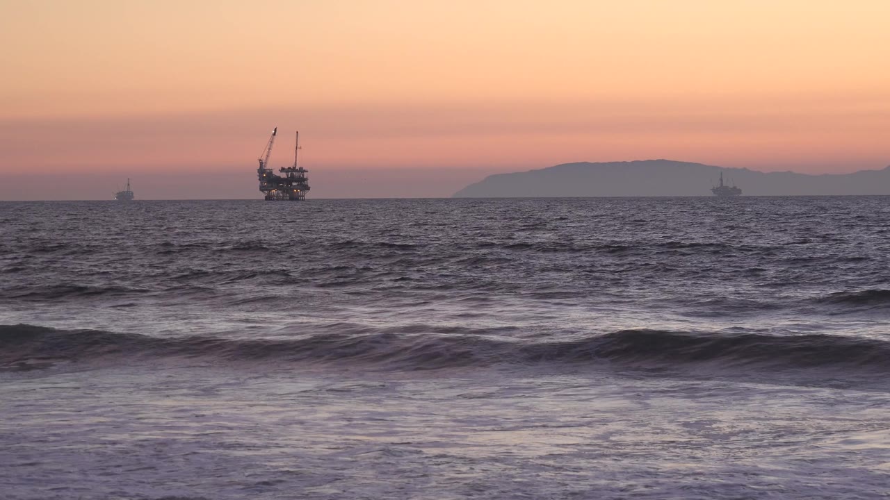 At Huntington Beach, dark midground water and low breaking waves sit below a layered sunset sky with offshore boats spread along the horizon, with no visible people.
