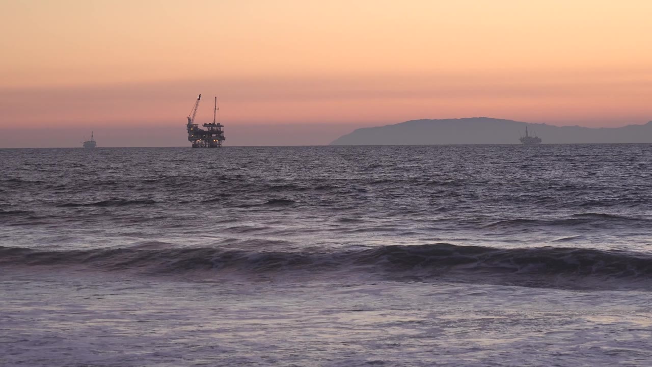 At Huntington Beach, a band of rough water crosses the foreground while distant boat silhouettes and faint land sit beneath the warm sky, with no visible people.