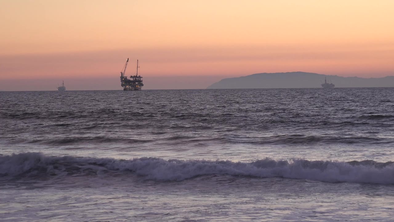 Huntington Beach surf rolls across the foreground while a larger offshore structure sits left of center near the horizon, with a muted landform behind it and no visible people.