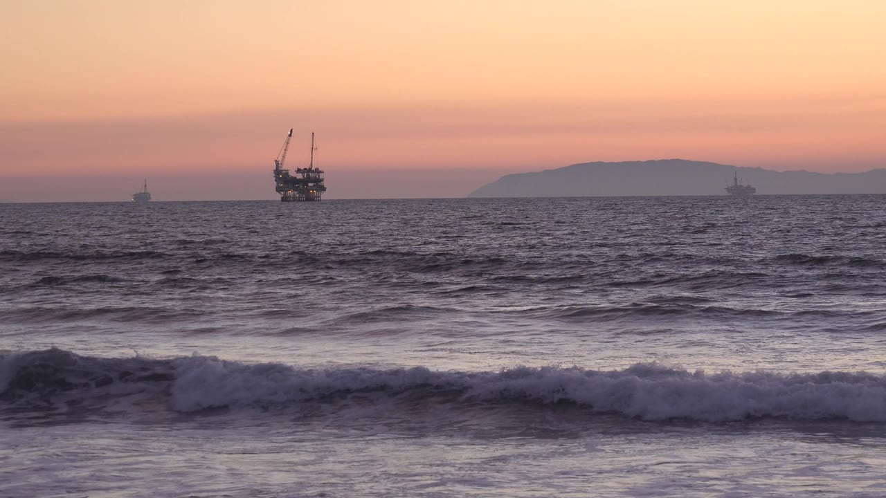 Huntington Beach ocean layers fill the frame with a darker wave band in front, small distant vessels spread along the horizon, a larger offshore structure near center, and no visible people.