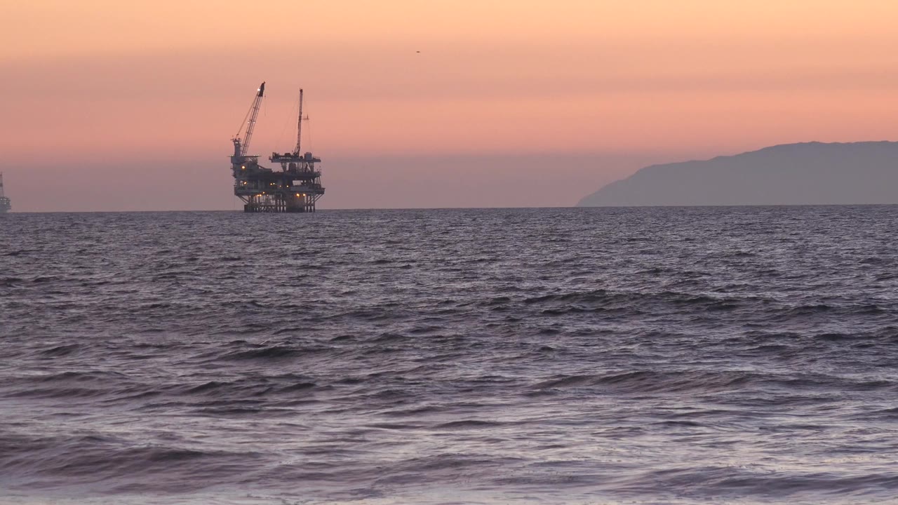 The offshore oil platform remains near the left horizon in Huntington Beach, California, with textured dark water in front, warm sunset sky above, and no visible people.