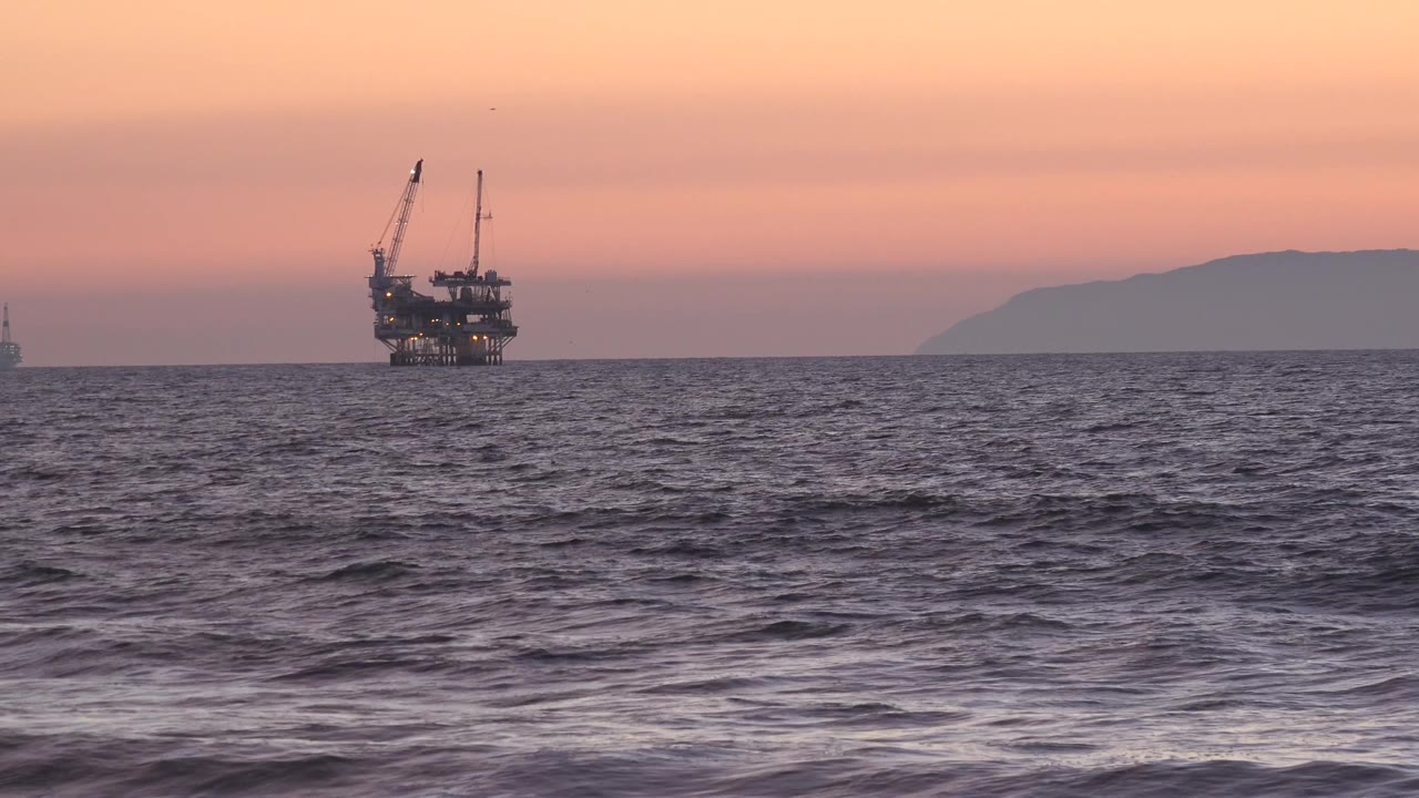 An offshore oil platform appears as a dark silhouette near the horizon at Huntington Beach, California, with the sea surface rougher in the foreground and no visible people.