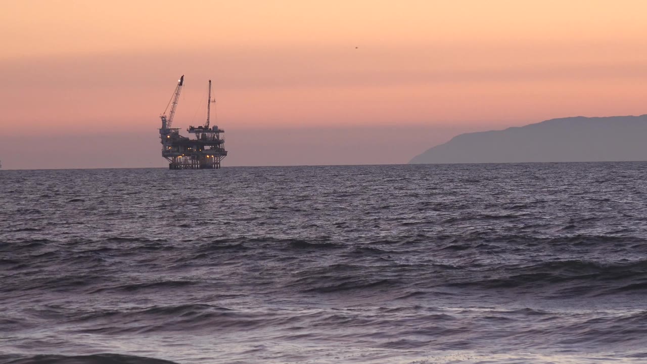 A distant metal platform with cranes sits near the left side of the Huntington Beach horizon while low waves cross the foreground and no visible people appear.