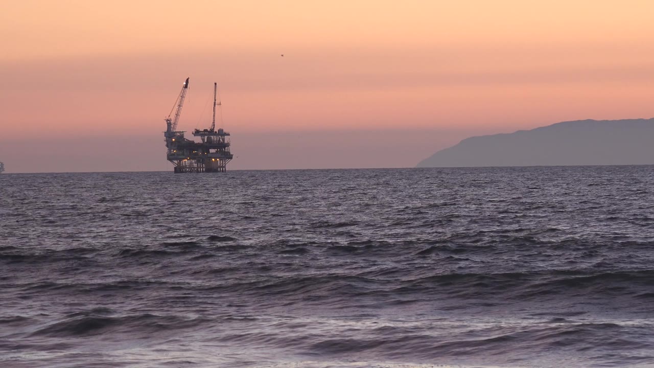 Sunset light washes the sky above the Huntington Beach water, with the industrial platform left of center and a hazy island or headland stretching along the right horizon, no visible people.