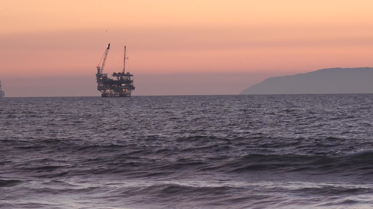 Low surf lines move across the Huntington Beach foreground while the offshore platform remains on the left horizon under muted orange and pink sky, with no visible people.